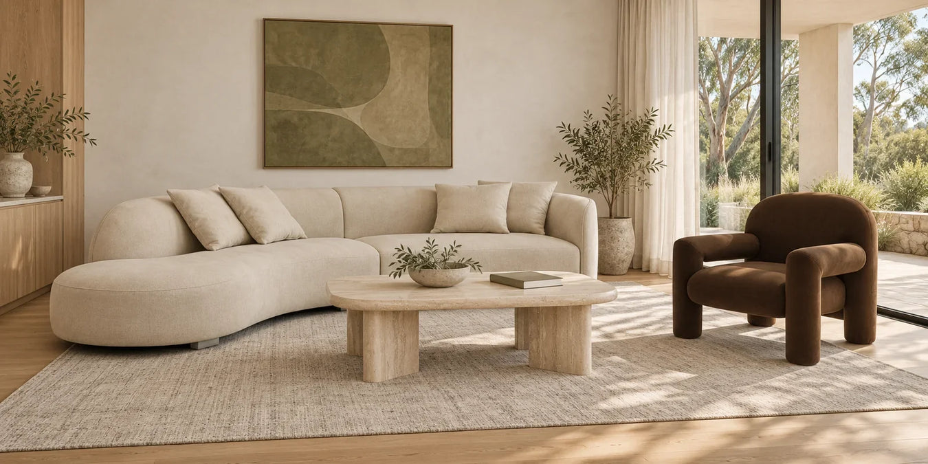 Curved ecru sofa, travertine coffee table and brown armchair on an oatmeal rug in a sunlit living room.