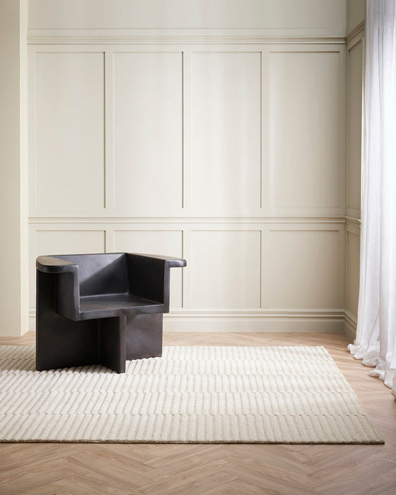 Anni Floor Rug in Oat, in a panelled room with a dark sculptural armchair on herringbone floors