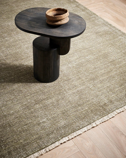 Corbett Floor Rug in Lichen, seen from above with a dark timber side table, showing the fringed edge
