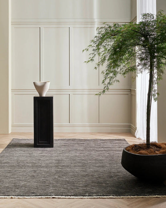 Corbett Floor Rug in Peat, in a panelled room with a black plinth and potted tree