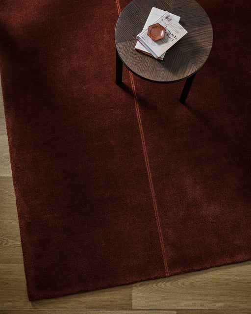 Bastion Floor Rug in Claret, seen from above with a round timber side table and stacked books