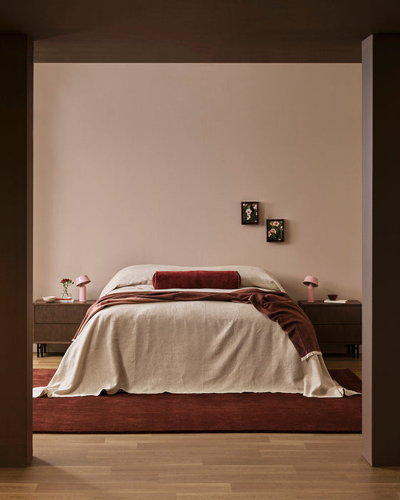 Bastion Floor Rug in Claret, under a bed with cream linen and timber bedsides in a bedroom