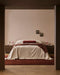 Bastion Floor Rug in Claret, under a bed with cream linen and timber bedsides in a bedroom