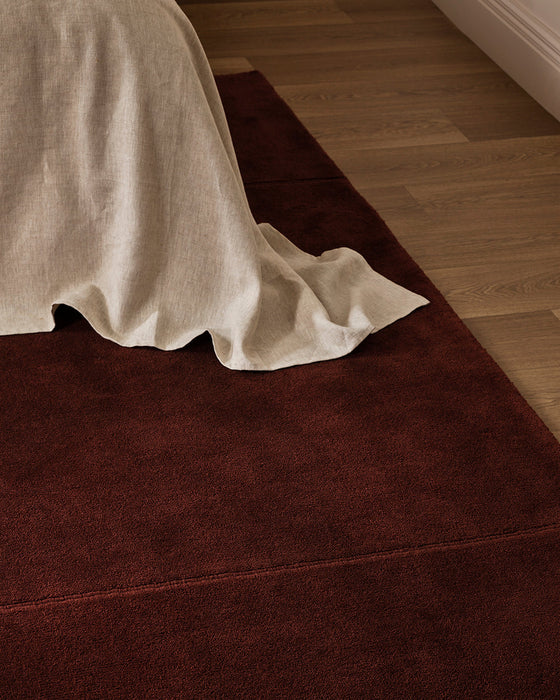 Bastion Floor Rug in Claret, close up at the foot of a bed draped in cream linen