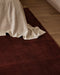 Bastion Floor Rug in Claret, close up at the foot of a bed draped in cream linen