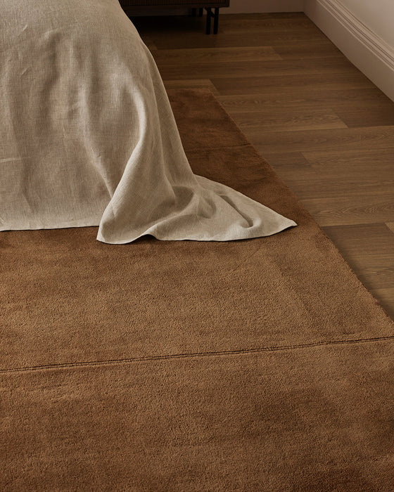 Bastion Floor Rug in Hazelnut, close up at the foot of a bed draped in cream linen