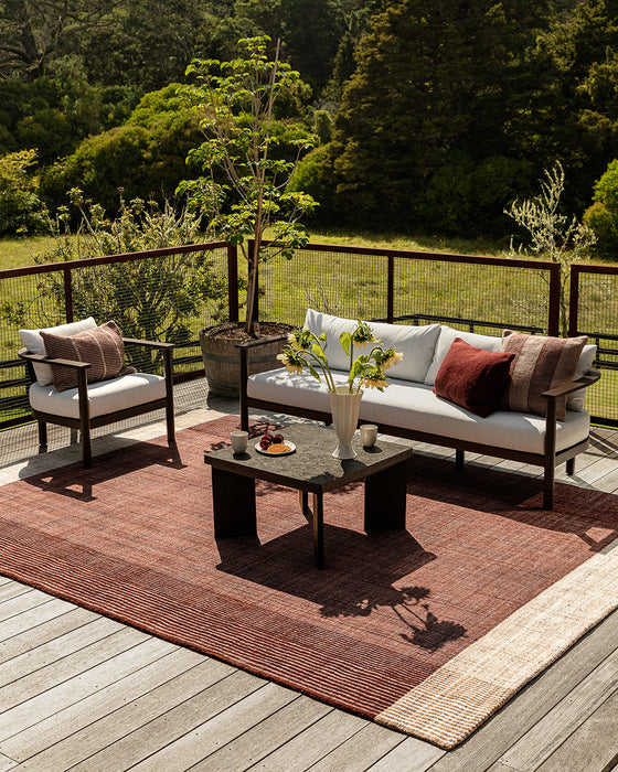 Bedrock Floor Rug in Claret, on a timber deck with an outdoor sofa, armchair and coffee table among trees