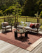 Bedrock Floor Rug in Claret, on a timber deck with an outdoor sofa, armchair and coffee table among trees