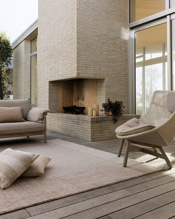Bedrock Floor Rug in Sand, on a covered deck beside an outdoor brick fireplace and woven lounge chair