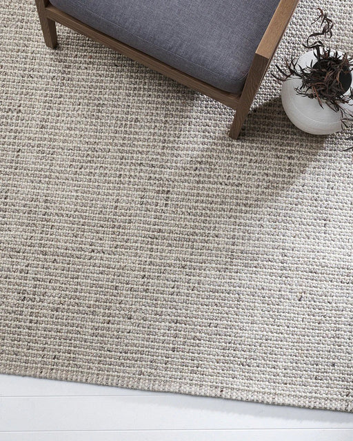 Kansas Floor Rug in Oyster/Natural, close view under a grey cushioned armchair, showing the chunky weave