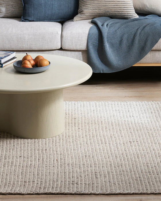 Kansas Floor Rug in Oyster/Natural, under a round cream coffee table in front of a grey sofa