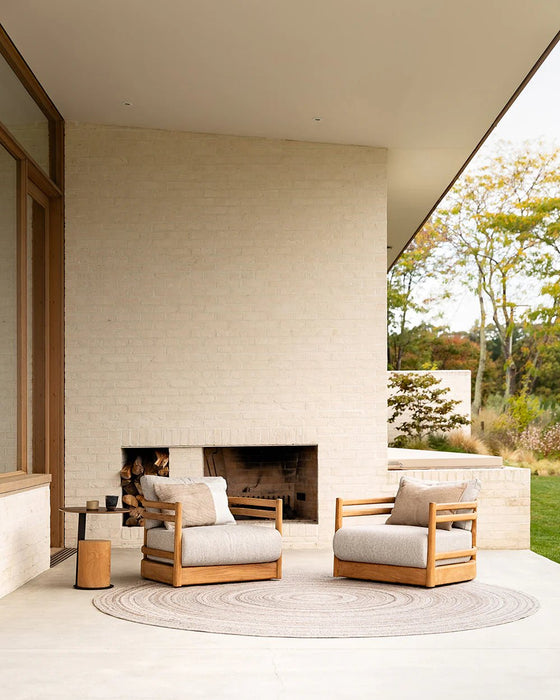 Palm Cove Round Floor Rug in Sand, on a covered terrace with two teak armchairs beside an outdoor fireplace