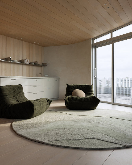 Edgewater Round Floor Rug in Pistachio, in a living room with dark green lounge chairs and a kitchen behind