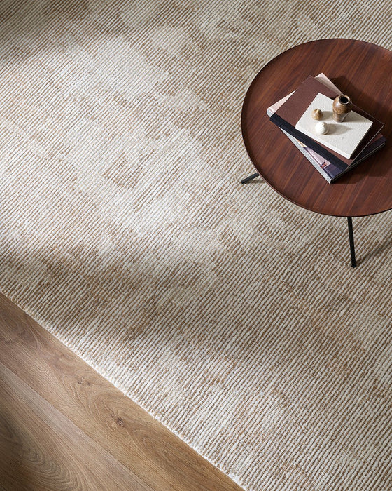 Beckett Floor Rug in Straw, seen from above beside a round timber side table, showing the abstract pattern