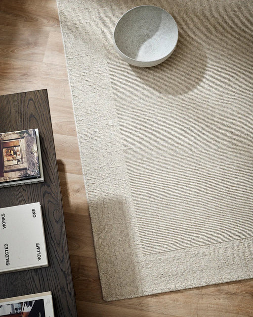 Vermont Floor Rug in Sand, seen from above beside a dark timber table with a ceramic bowl