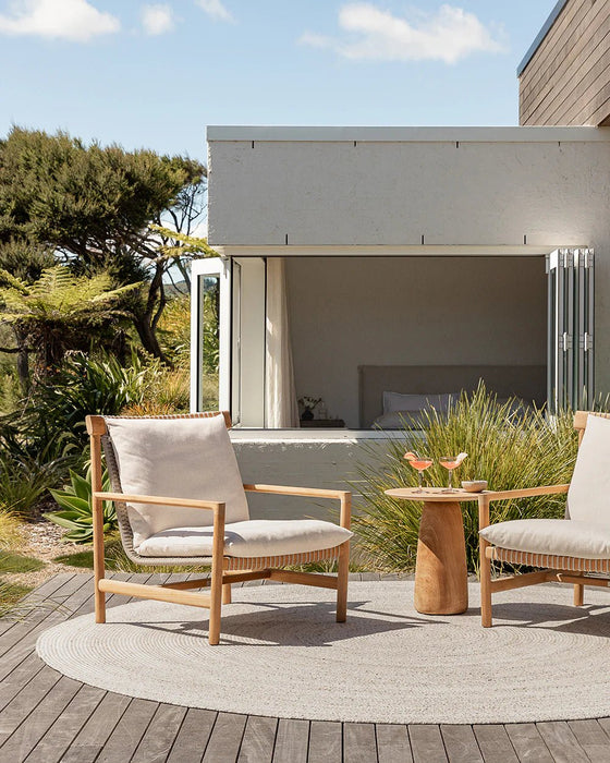 Mornington Round Floor Rug in Pale Sand, on a timber deck beside a white house with two outdoor armchairs