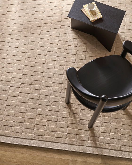 Hugo Floor Rug in Almond, seen from above with a black chair and side table, showing the raised block pattern