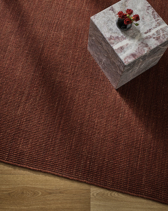 Linden Floor Rug in Brick, seen from above with a marble side table holding red flowers