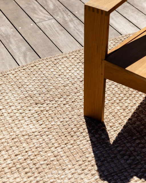 Pelorus Floor Rug in Sand, close up on a timber deck under a teak chair, showing the woven texture