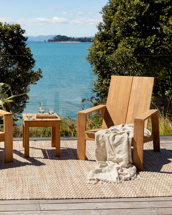 Pelorus Floor Rug in Sand, on a timber deck overlooking the sea with a teak lounge chair and throw