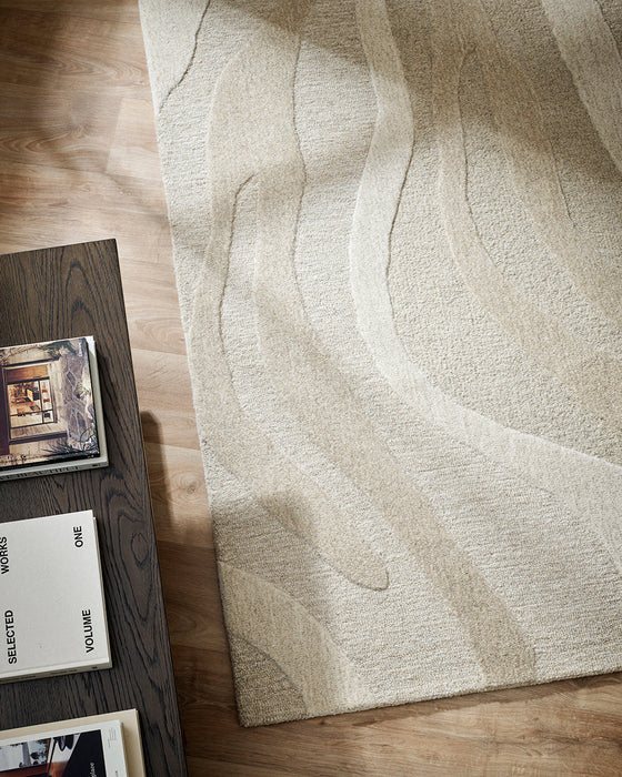 Shoreline Floor Rug in Sand, seen from above beside a dark timber table, showing the carved waves