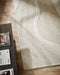 Shoreline Floor Rug in Sand, seen from above beside a dark timber table, showing the carved waves