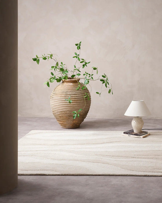 Shoreline Floor Rug in Sand, in a minimal room with a large clay vase and table lamp