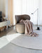 Tairua Round Floor Rug in Natural Straw, under a curved armchair with a pink throw beside a floor lamp
