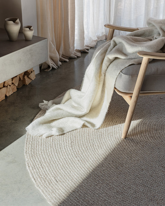 Tairua Round Floor Rug in Natural Straw, close up under a timber armchair with a cream throw on a concrete floor