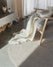 Tairua Round Floor Rug in Natural Straw, close up under a timber armchair with a cream throw on a concrete floor