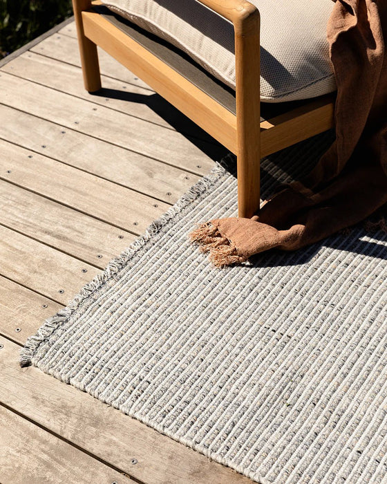 Rhodes Floor Rug in Slate, close up on a timber deck under a teak chair with a rust throw