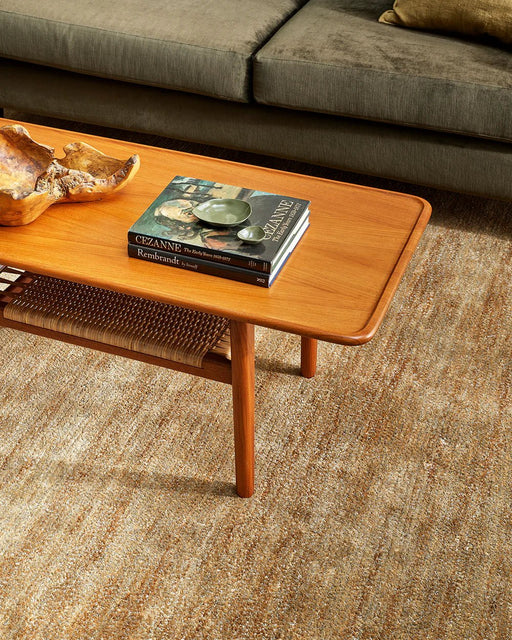 Fairbanks Floor Rug in Sand, under a timber coffee table with a rattan shelf beside an olive sofa
