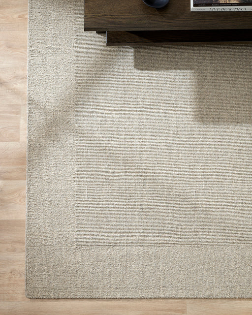 Vermont Floor Rug in Driftwood, seen from above under a dark timber coffee table, showing the bordered weave